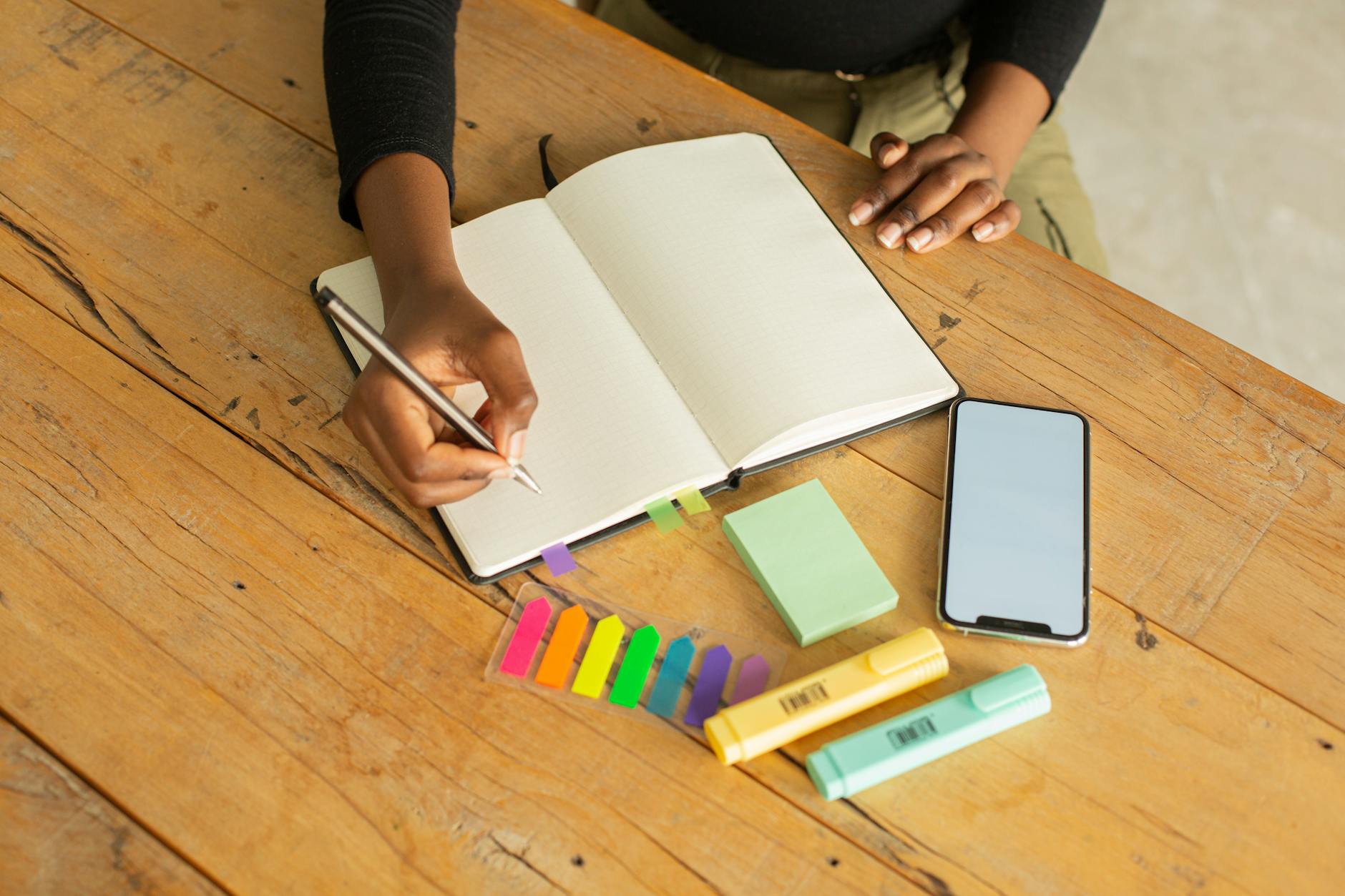 crop unrecognizable black woman writing in notebook and using smartphone